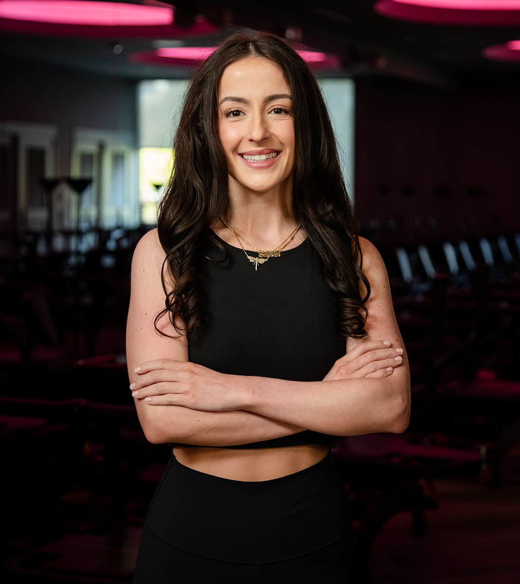 sydneigh-fitness-instructor-portrait-with-arms-crossed-in-red-lit-studio.tight-dallas. Sydneigh, fitness instructor, standing with arms crossed and smiling in a dimly lit Lagree studio.