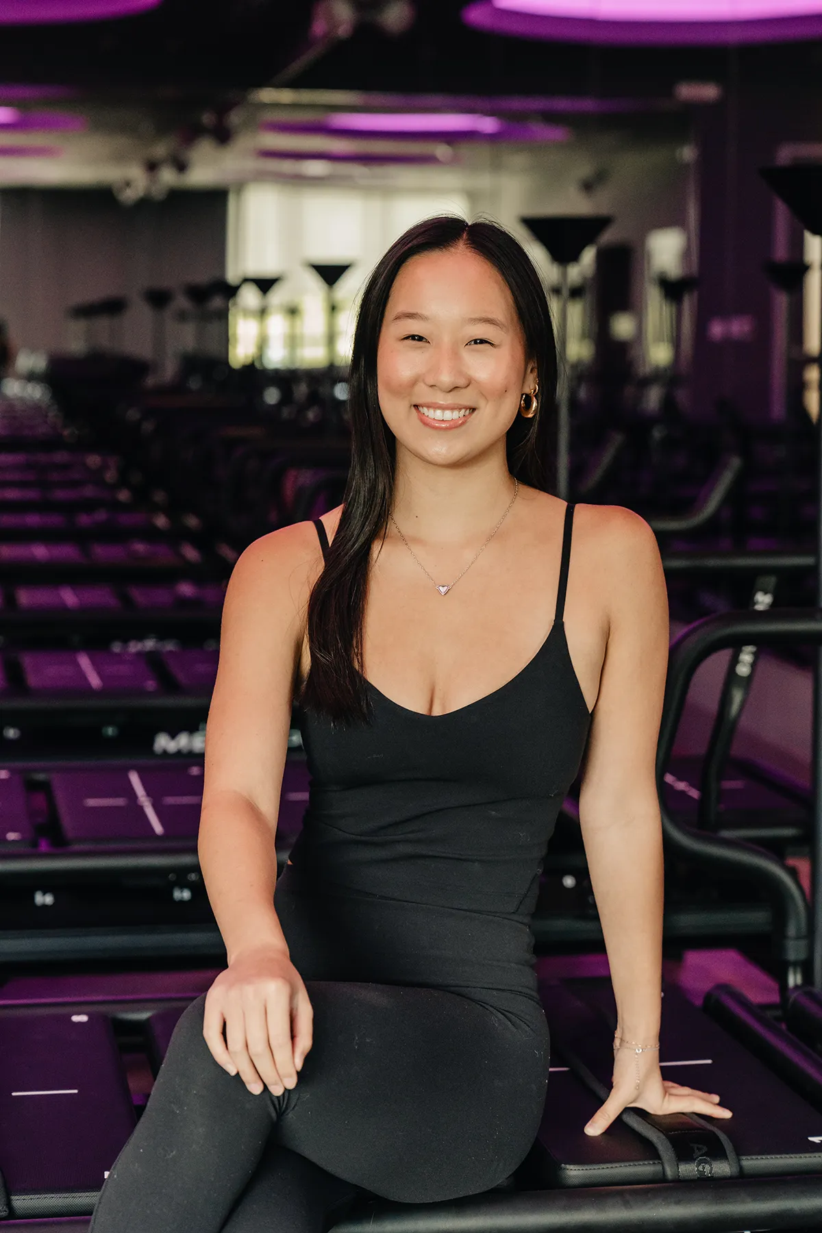 nina-lagree-instructor-seated-portrait-in-purple-lit-megaformer-studio.tight-dallas. Nina, Lagree instructor, smiling while seated on a Megaformer in a purple-lit studio.