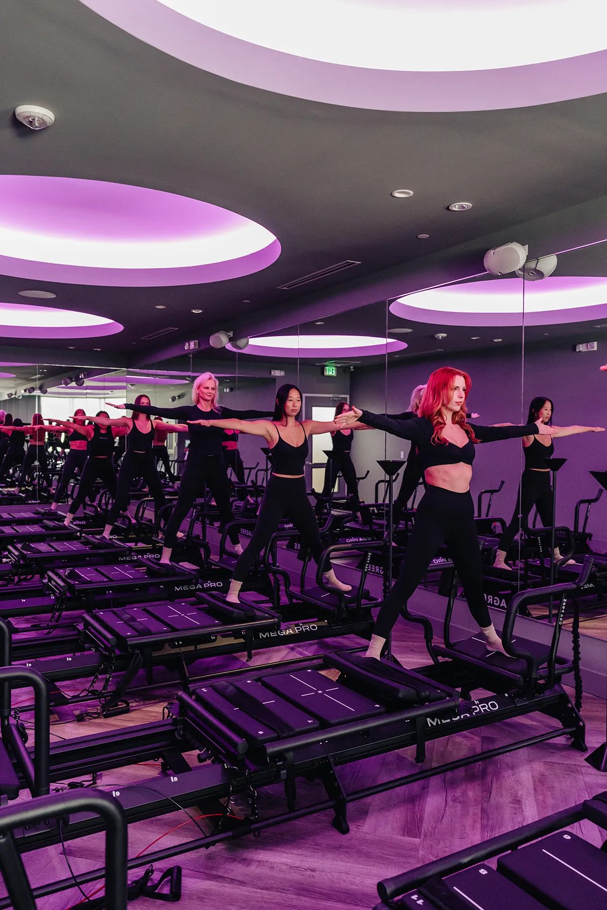 group-lagree-class-standing-exercise-on-megaformer-machines-with-purple-lighting.tight-dallas. Group fitness class performing standing exercises on Megaformer machines under purple lighting.