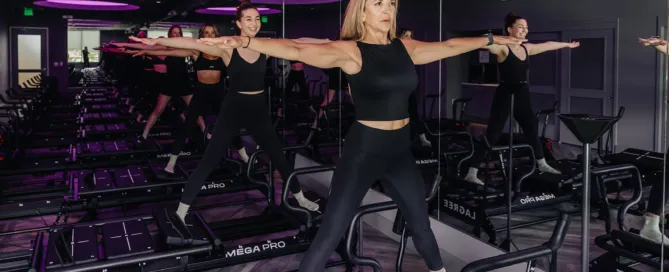 Group of women in black activewear performing a wide-stance arm extension on Lagree Mega Pro machines in a purple-lit boutique fitness studio.