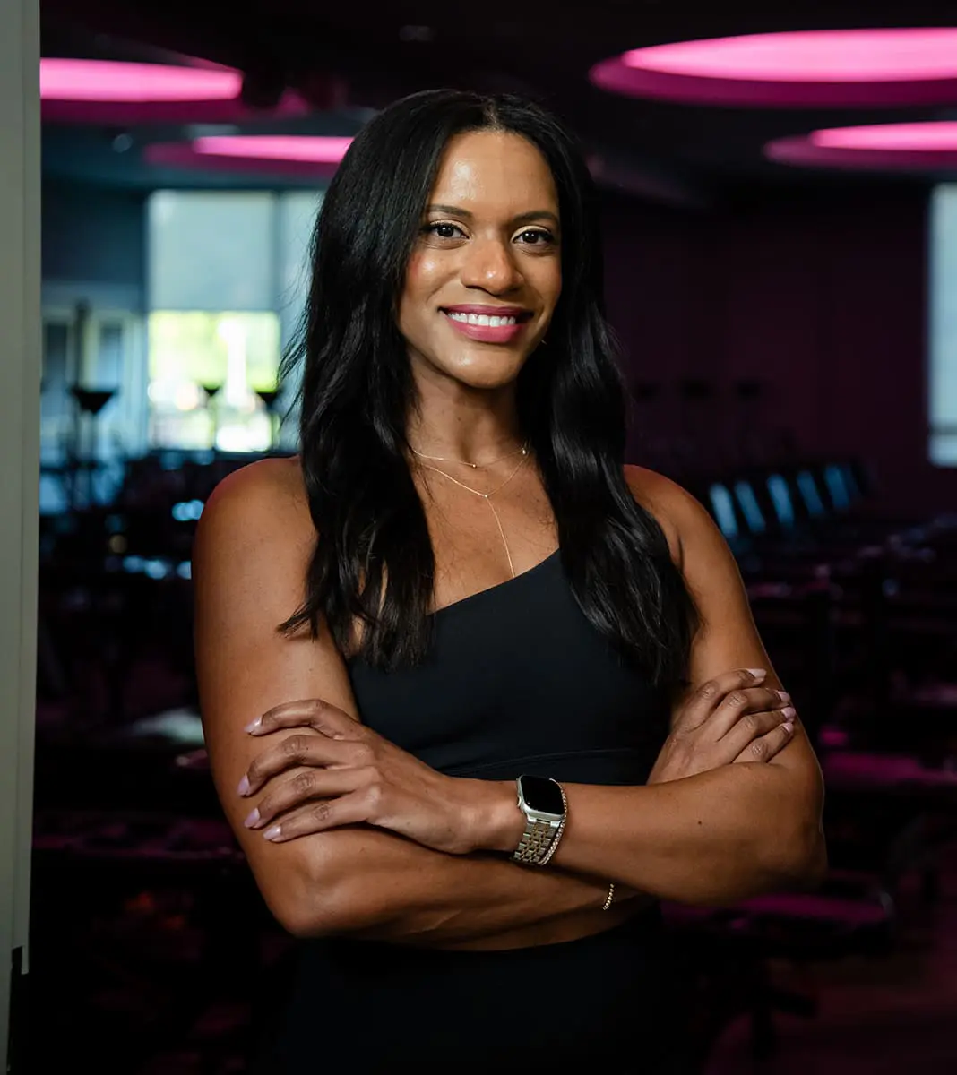 kim-lagree-instructor-portrait-with-arms-crossed-in-pink-lit-studio.tight-dallas. Kim, Lagree instructor, smiling with arms crossed under pink studio lights.