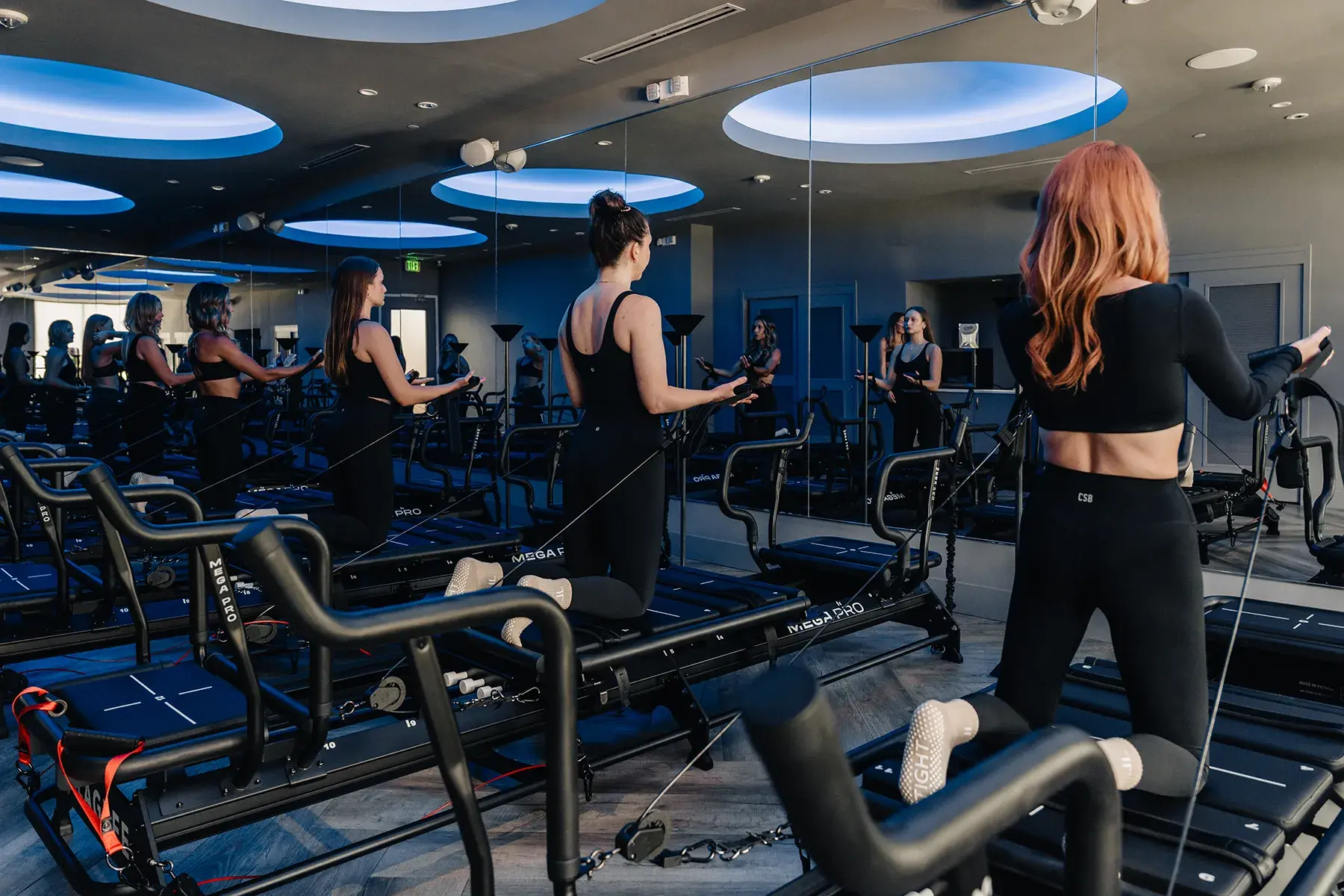 lagree-class-kneeling-resistance-exercise-on-megaformer-machines-in-modern-studio.tight-dallas. Women performing resistance exercises on Megaformer machines in a mirrored fitness studio.