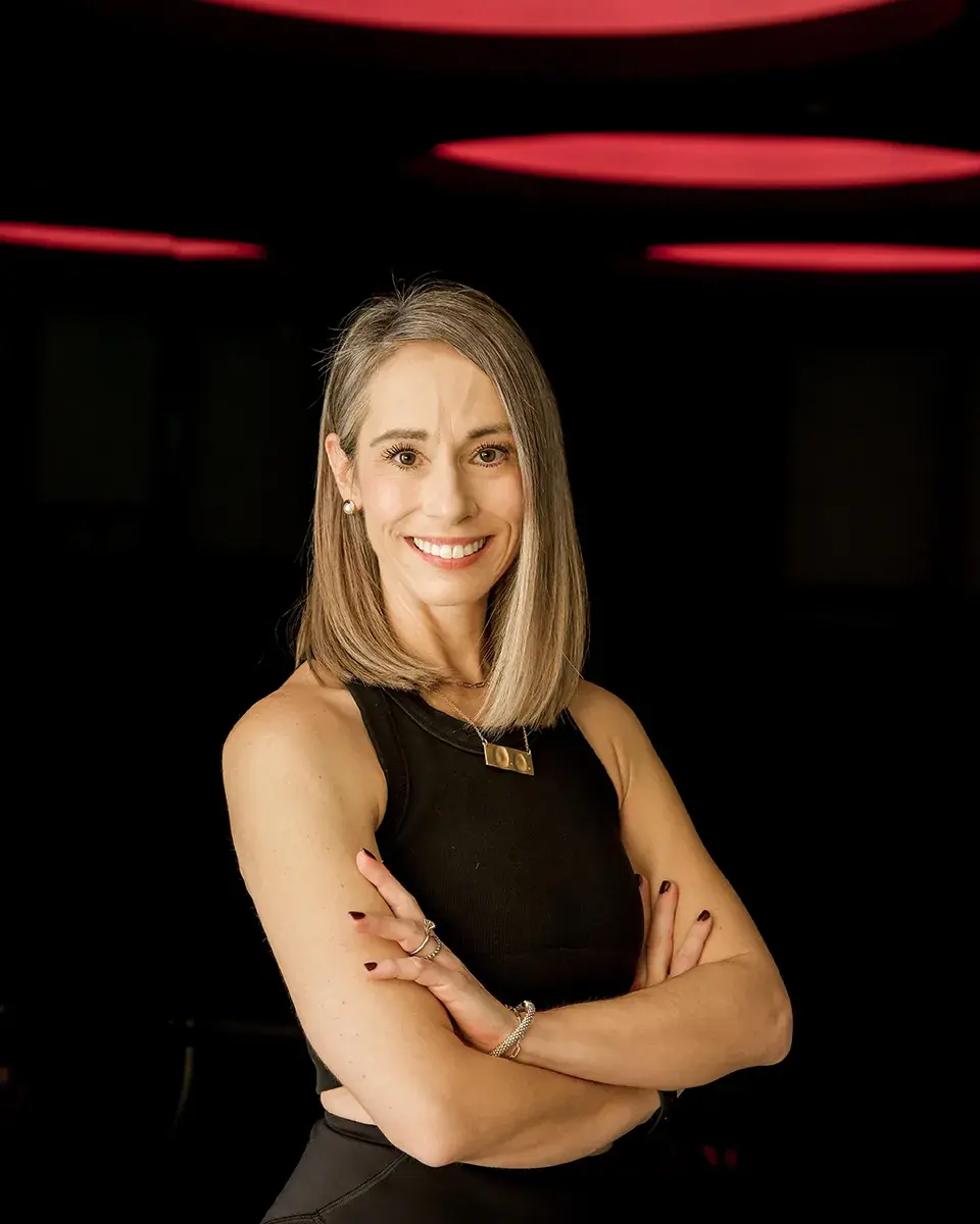 nicole-lagree-instructor-portrait-with-arms-crossed-in-dark-studio-lighting.tight-dallas. Nicole, Lagree instructor, smiling with arms crossed against a dark studio backdrop.