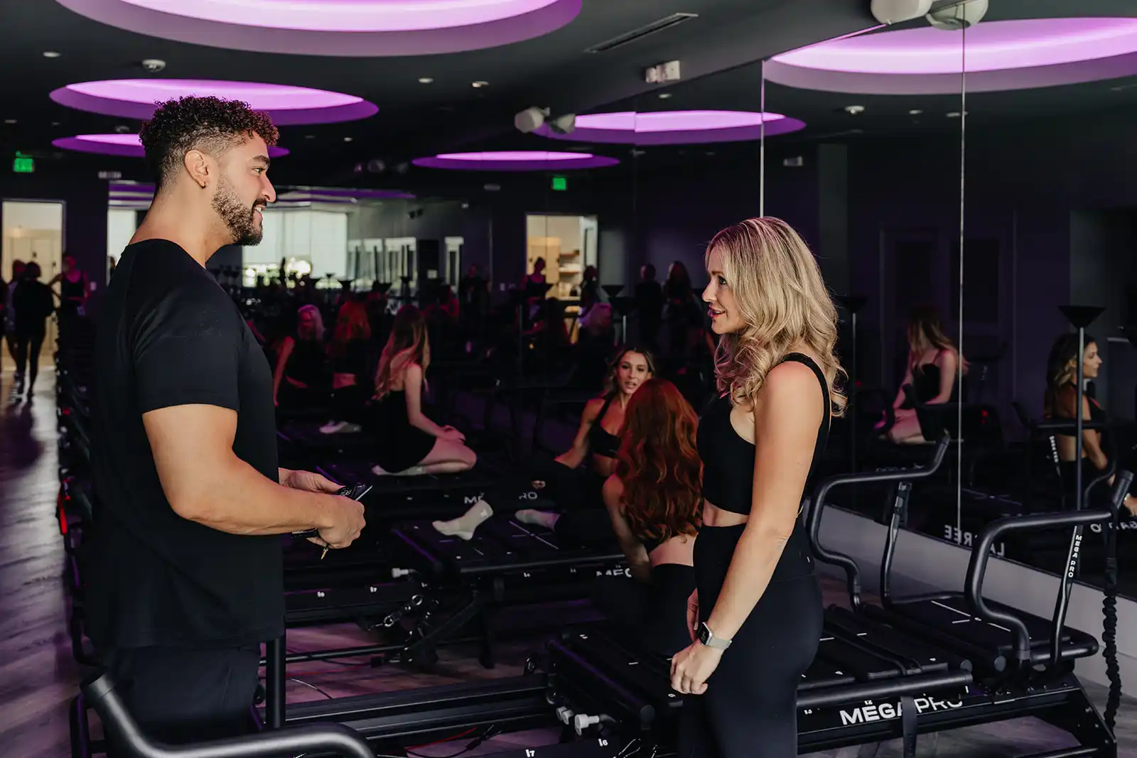 instructors-talking-megaformer-lagree-studio-tight-dallas. Two TIGHT Dallas instructors chatting between Megaformer machines in a purple-lit Lagree studio