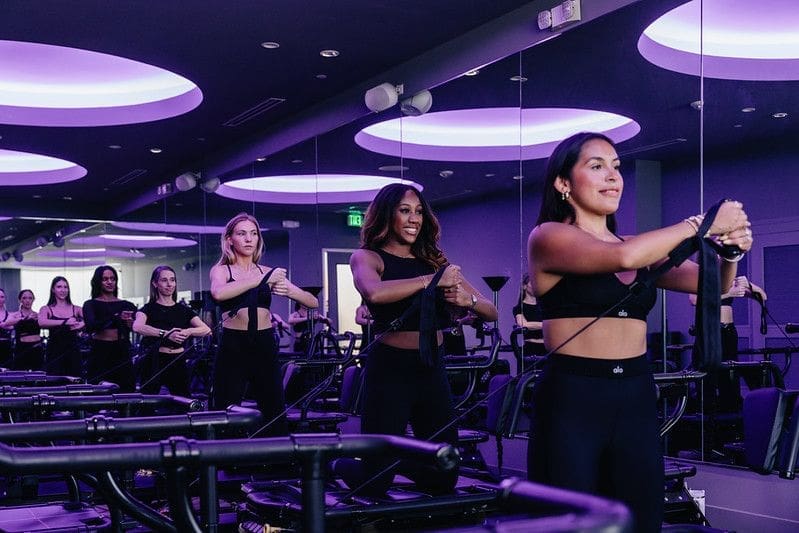 T0NFnl_A Women participating in a small group Lagree fitness class at TIGHT Dallas, using Megaformer machines in a modern studio with purple lighting.