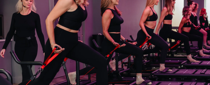 Group of women performing Lagree workout on Megaformer machines with resistance bands, guided by an instructor in a fitness studio.