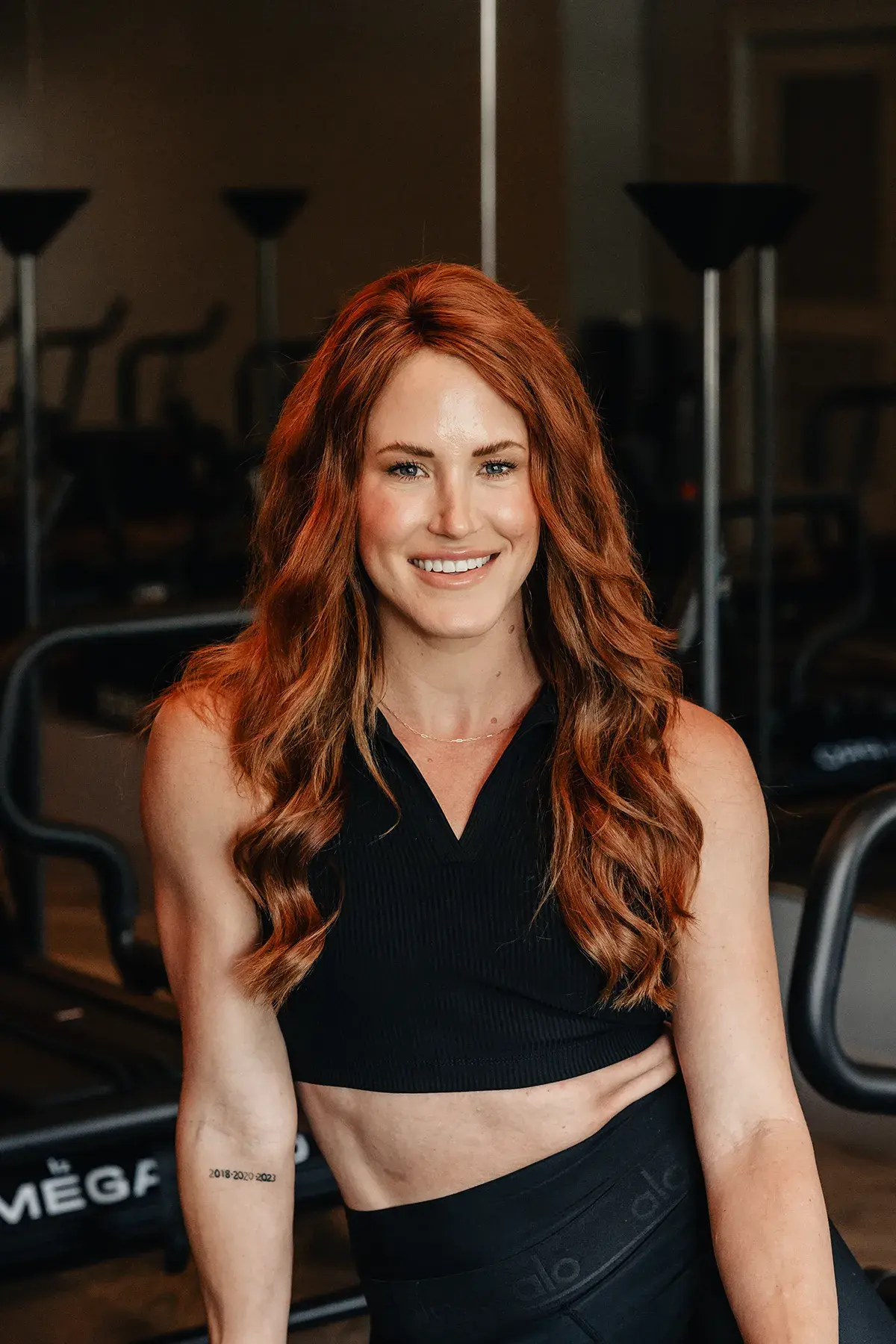Sarie, Lagree instructor, smiling with long red hair while leaning slightly forward in a Megaformer studio.